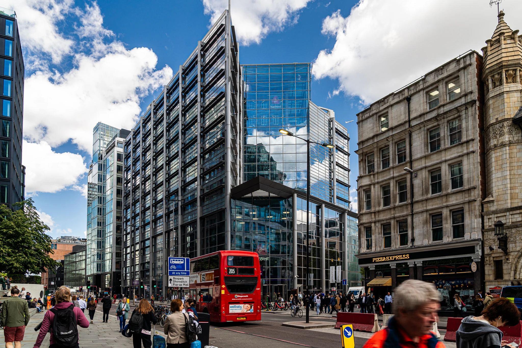 Czerwony piętrowy autobus przejeżdżający obok nowoczesnych szklanych budynków i tradycyjnej zabudowy w pobliżu Liverpool Street w Londynie, w słoneczny dzień z przechodniami.