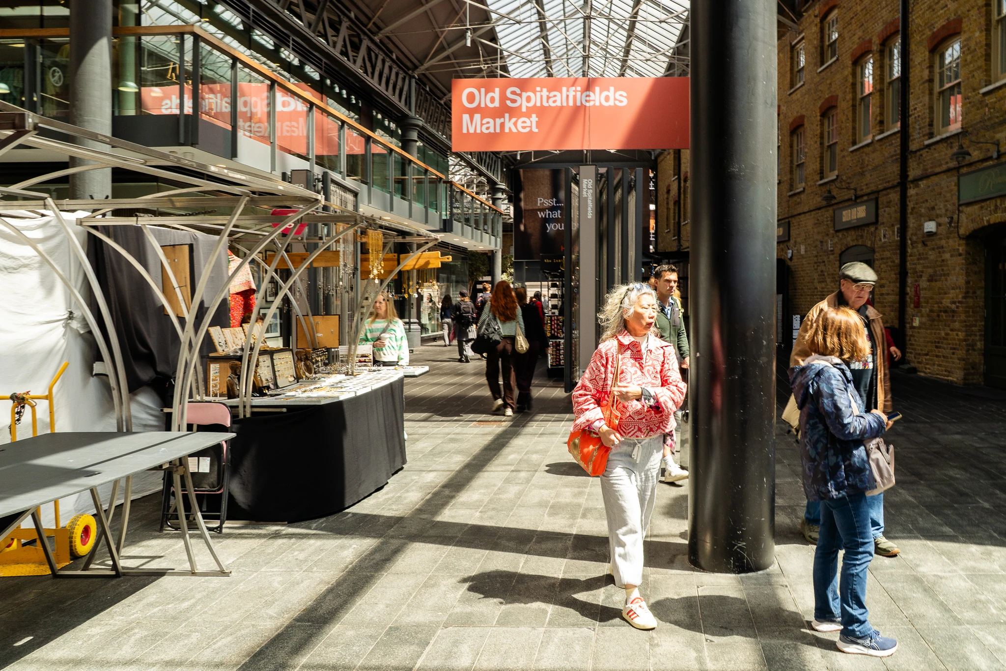 Ludzie przechadzający się po Old Spitalfields Market w Londynie, obok stoisk z biżuterią i innymi produktami, w jasnym świetle słonecznym.