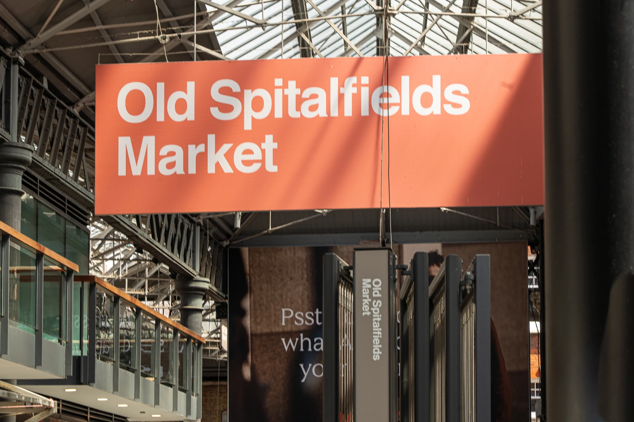 Duży, pomarańczowy szyld z napisem „Old Spitalfields Market” zawieszony w zabytkowym wnętrzu targowiska w Londynie.