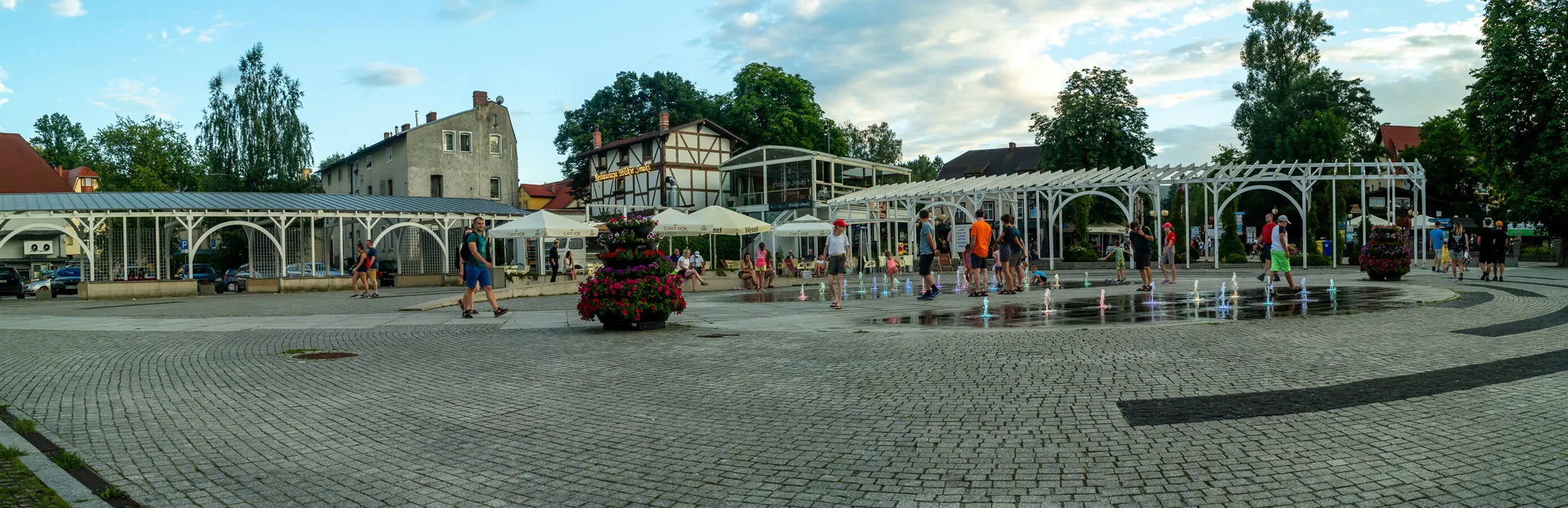 Panorama Małego Rynku w Polanicy Zdrój z podświetloną kolorowymi lampami fontanną i białą pergolą pełną turystów
